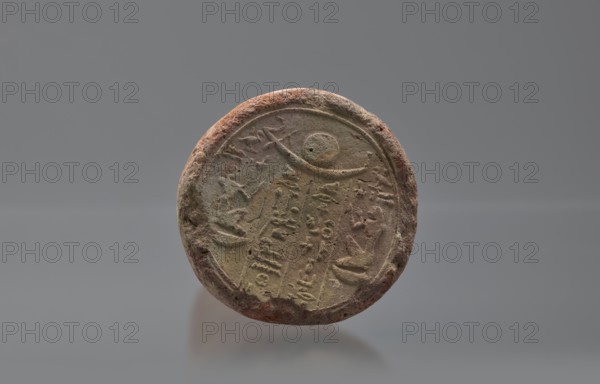 Basa's funeral cone, created by Basa ca. 664–610 BC. The hieroglyphic inscription on the base runs in two vertical columns. On either side a figure facing the inscription, kneeling on the neb sign, with hands raised in prayer. Above the inscription is a representation of a solar barque. Boat and solar disc iconography; hieroglyphs. Scen.Gal.Eg. Davies-Macadam, Corpus No. 602. Date: 664–610 BC; Late Period, 26th Dynasty reign of Psammetik I. Provenance: Western Thebes tomb 389 on Asasif. Cone-shaped funerary cone; height 9 cm, width 23 cm, weight 1.072 kg., A round, weathered object with a rough, uneven rim sits against a gray background; the object shows textured surfaces and carved or impressed shapes and markings across its face, with a raised circular border and a crescent-like curved line near the top, and the surface displays a mix of earthy tones including brown, reddish-brown, beige, and muted green.