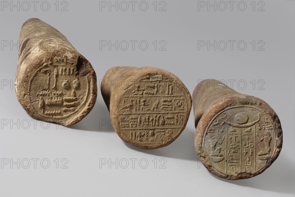 Basa's funeral cone, maker, Basa ca. 664–610 BC. Cone-shaped funerary cone with hieroglyphic inscription on the base in two vertical columns. On either side a kneeling figure facing the inscription, kneeling on the neb sign with hands raised in prayer. Above the inscription is a representation of a solar barque boat and a solar disc. Late Period, 26th Dynasty reign of Psammetich I, ca. 664–610 BC. From Western Thebes tomb 389 on Asasif. Height 9 cm; width 23 cm; weight 1.072 kg. Davies-Macadam, Corpus No. 602., A group of three cylindrical stone objects lie on a pale gray surface against a light gray background, each showing a round carved end with raised designs. The left cylinder is elongated and tan with rough brown and beige textures and a circular carved face that includes concentric rings and raised motifs; it casts a soft shadow to its right. The middle cylinder is shorter and squat, colored warm brown and sandy beige with a worn circular end carved with linear raised patterns a...