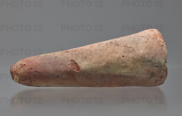 Basa's funeral cone, Basa ca. 664–610 BC. Cone-shaped funerary cone, height 9 cm, width 23 cm, weight 1.072 kg. Late Period, 26th Dynasty reign of Psametik I, Western Thebes tomb 389 on Asasif. The hieroglyphic inscription on the base runs in two vertical columns. On either side a figure facing the inscription, kneeling on the neb sign, with hands raised in prayer. Above the inscription is a representation of a solar barque. Iconography: boat, solar disc; contains hieroglyphs. Davies-Macadam, Corpus No. 602., A single elongated, tapered object lies horizontally against a neutral gray background, showing a rough, pitted surface with a mix of muted reddish, brown, beige, and gray tones and areas of greenish discoloration; the shape is thicker and rounded on one end and narrows to a blunter tip on the other, with visible small holes, scratches, and uneven texture across its surface and a faint soft reflection beneath it.