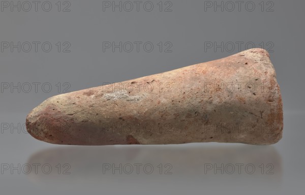 Basa's funeral cone, creator, Basa ca. 664–610 BC. Cone-shaped funerary cone, height 9 cm, width 23 cm, weight 1.072 kg. Late Period, 26th Dynasty reign of Psammetik I, Western Thebes tomb 389 on Asasif. The hieroglyphic inscription on the base runs in two vertical columns. On either side a figure faces the inscription, kneeling on the neb sign with hands raised in prayer. Above the inscription is a representation of a solar barque. Iconography: boat, solar disc; contains hieroglyphs. Davies-Macadam, Corpus No. 602., A single elongated, conical object rests horizontally against a smooth gray background with a soft reflection beneath it; the object has a rough, pitted surface showing a mix of earthy colors including tan, beige, reddish-brown, pinkish hues, and patches of light gray, with some darker spots and mottled texture across its surface.