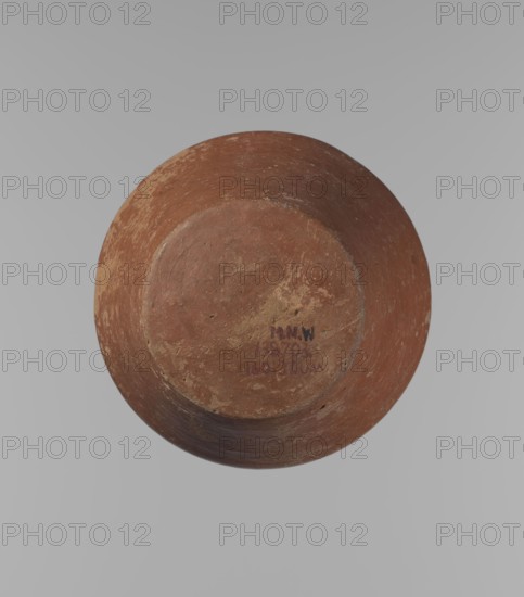 Vessel, Egyptian pottery, ceramics, clay vessel. A conical vessel, widest at approximately 3/4 of its height. The shoulders are quite wide, almost horizontal—slightly sloping. The neck is wide, short, funnel-shaped, widening towards the spout. The spout is wide, flat in the upper part, slightly rounded at the bottom, slightly asymmetrical. The bottom is slightly convex, asymmetrical. The surface of the vessel is even. The lower part of the vessel has been worked to approximately 2/3 of its height with a knife or similar tool. The outer surface of the vessel and the inner surface of the rim and neck are covered with a layer of dark red engobe. Vessel color: dark red. Vessel complete. Surface eroded in many places, numerous clay chips on the rim. ca. 2686–ca. 2181 BC; Old Kingdom, Edfu; vessel; height 16.7 cm, diameter 9.5 cm rim, diameter 13 cm body, diameter 8.1 cm bottom., A round, shallow ceramic bowl viewed from above showing its underside, with a worn, textured surface in reddish-b...