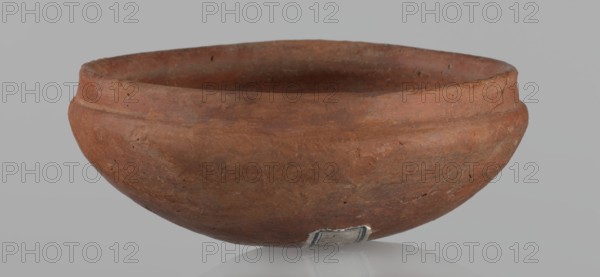 Medum-type bowl, A small Medum-type bowl with characteristic carination under the rim. Rounded bottom. Arched walls, gently breaking towards the center of the vessel around 2/3 of its height. Straight, perpendicular rim. Narrow, sloping shoulders. The diameter of the rim is significantly smaller than the diameter of the vessel's arms. Clay probably NB2. Surface leveled and covered with a layer of engobe, then polished. Vessel color: red. c. 2686–c. 2181 BC; Old Kingdom, Edfu. Vessel; height 4.5 cm, diameter 11 cm rim, diameter 11.6 cm arms., A rounded shallow bowl with a matte, earthy surface in shades of reddish-brown and terracotta sits against a pale gray background, showing a slightly uneven rim and a subtle raised band around its upper exterior; the interior appears darker with similar reddish-brown tones, and the outer surface has a few small pits and rough patches, along with a small area near the base that shows a fragment of a rectangular label with worn edges in muted blue an...