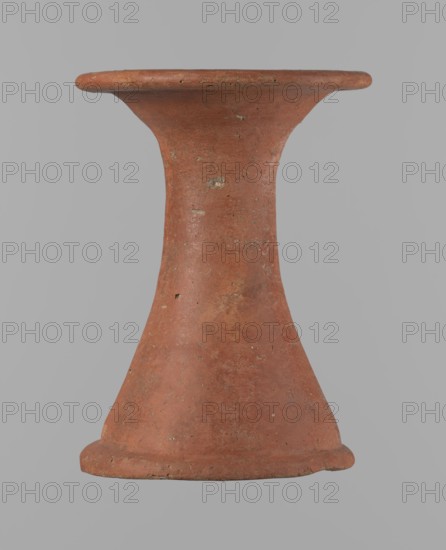 Bowl stand, Base for a vessel with an almost equal diameter of both rims. Both rims are irregular, with a slightly wavy line. The center of the base is empty. The narrowest part of the base is approximately 2/3 of the height of the entire vessel. Made of Nile clay NB2. The surface is leveled and covered with an engobe, then polished. Red in color. Complete, very well preserved. Set with bowl 138673. ca. 2494–ca. 2181 BC; Old Kingdom, 5th–6th Dynasty, Edfu. Vessel; height 13.7 cm; diameter 10.2 cm rim; diameter 9.3 cm bottom., An upright hourglass-shaped clay object with a flared rim and base, showing a weathered surface with small chips and rough patches, in shades of reddish-brown and terracotta against a flat gray background.