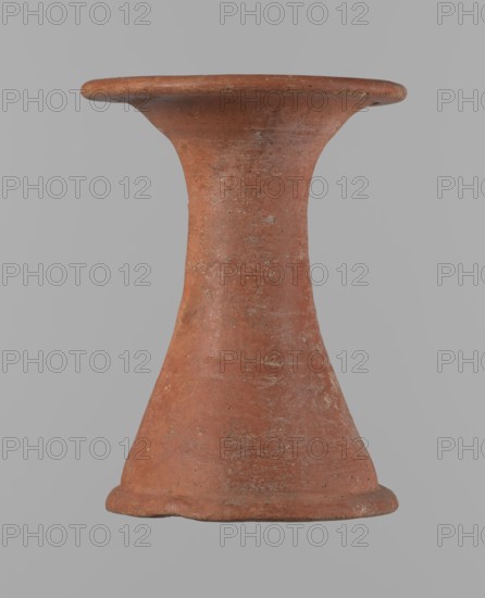 Bowl stand;, Base for a vessel with an almost equal diameter of both rims. Both rims are irregular with a slightly wavy line. The center of the base is hollow. The narrowest part of the base is approximately 2/3 of the height of the entire vessel. Made of Nile clay NB2. Surface leveled, covered with an engobe, then polished. Red color. Complete, very well preserved. Set with bowl 138673. Circa 2494–2181 BC; Old Kingdom, 5th–6th Dynasty, Edfu. Vessel; height 13.7 cm; diameter 10.2 cm rim, 9.3 cm bottom., A single terracotta object shaped like an hourglass with a flared rim at the top and a rounded base sits against a plain light gray background; the surface of the object is textured with weathered spots and small patches of lighter discoloration, and the predominant color is a muted reddish-brown with traces of pale gray and occasional darker brown specks.
