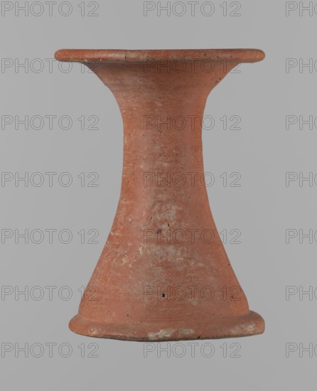 Bowl stand. Base for a vessel with an almost equal diameter of both rims. Both rims are irregular, with a slightly wavy line. The center of the base is empty. The narrowest part of the base is approximately 2/3 of the height of the entire vessel. Made of Nile clay NB2. The surface is leveled and covered with an engobe, then polished. Vessel color: red. Complete, very well preserved. Set with bowl 138673. Ca. 2494–ca. 2181 BC; Old Kingdom, 5th–6th Dynasty, Edfu. Vessel; height 13.7 cm; diameter 10.2 cm rim; diameter 9.3 cm bottom., A terracotta object with a flared top and bottom connected by a narrow waist, showing a weathered reddish-brown surface with patches of lighter beige and gray discoloration, small dark pitted marks, and irregular rough texture against a plain light gray background.