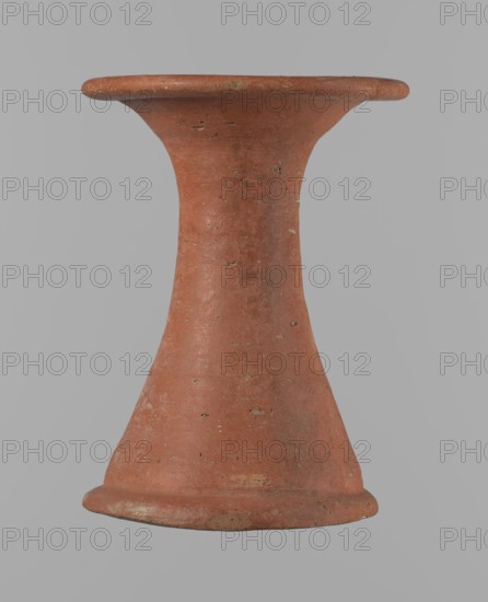 Bowl stand, Base for a vessel with an almost equal diameter of both rims. Both rims are irregular, with a slightly wavy line. The center of the base is empty. The narrowest part of the base is approximately 2/3 of the height of the entire vessel. Vessel made of Nile clay NB2. The surface is leveled and covered with an engobe, then polished. Vessel color: red. Complete vessel, very well preserved. Set with bowl 138673. ca. 2494–ca. 2181 BC; Old Kingdom, 5th–6th Dynasty, Edfu. Vessel, height 13.7 cm; diameter 10.2 cm rim; diameter 9.3 cm bottom., A single terracotta object shaped like an hourglass or spool, with a flared rim at the top and a flared base at the bottom, shown against a plain light gray background. The object is primarily reddish-brown with small patches of lighter beige and darker brown where the surface shows wear or texture. Its surface has a rough, slightly pitted texture with scattered small irregularities and tiny chips. The silhouette tapers inward at the midsection ...