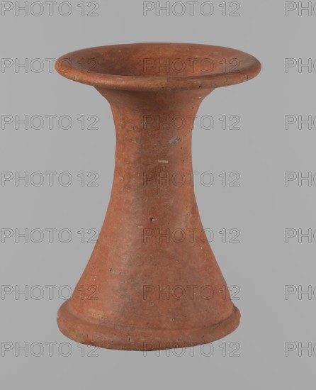 Bowl stand, Base for a vessel with an almost equal diameter of both rims; both rims are irregular with a slightly wavy line. The center of the base is empty. The narrowest part of the base is approximately 2/3 of the height of the entire vessel. Made of Nile clay NB2. Surface leveled, covered with an engobe, then polished. Color: red. Complete, very well preserved. Set with bowl 138673. ca. 2494–ca. 2181 BC; Old Kingdom, 5th–6th Dynasty, Edfu. Vessel; height 13.7 cm; diameter 10.2 cm rim, 9.3 cm bottom., A single terracotta object with a flared rim and a wider base, shaped like an hourglass with a narrow central neck; its surface shows small rough patches and spots of wear, and the visible colors include reddish-brown, muted orange, and hints of gray against a plain light gray background.