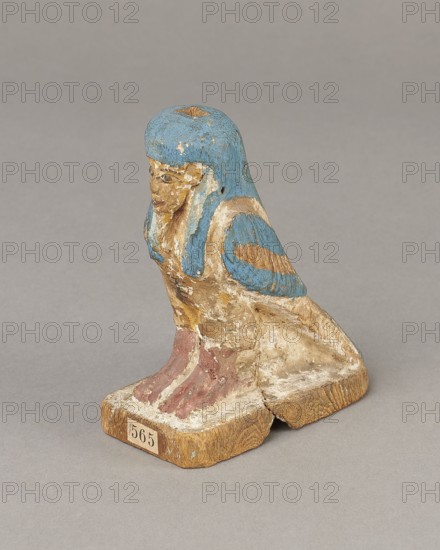 Figurine of the Soul of Ba, Ba mythology, birds, bird iconography, djed pillars iconography, wigs, Soul personification, figurines, beards facial hair, feathers. The figurine has carefully carved and painted facial details and a reproduced arrangement of feathers on the wings. On the head is a tripartite wig painted blue. On the chest below the wig is a depiction of a djed pillar. Straps supporting a false beard are painted along the cheeks. The figurine's disproportionately large feet rest on a rectangular base. 664–332 BC, Late Period, Egypt; figurine; height 11 cm, width 9.5 cm, depth 4.5 cm., A small carved figure resembling a seated creature with a humanlike face and a birdlike body sits on a rectangular base; the figure has a rounded blue headdress with worn areas, a face and body showing faded tan and beige paint with traces of white, blue painted wings with tan patches, and reddish-pink painted feet on a wood-toned base that shows cracks and aged texture.