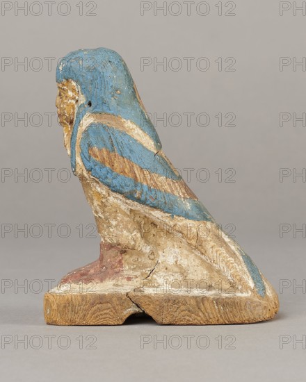 Figurine of the Soul of Ba, Ba mythology; birds, bird iconography; djed pillars iconography; wigs; Soul personification; figurines; beards facial hair; feathers. The figurine has carefully carved and painted facial details and a reproduced arrangement of feathers on the wings. On the head is a tripartite wig painted blue. On the chest below the wig is a depiction of a djed pillar. Straps supporting a false beard are painted along the cheeks. The figurine's disproportionately large feet rest on a rectangular base. 664–332 BC, Late Period, Egypt. Medium: figurine. Dimensions: height 11 cm, width 9.5 cm, depth 4.5 cm., A small carved figure resembling a perched bird with a humanlike face, shown in profile, painted in shades of blue, beige, white, brown, and a muted red; the figure sits on a rectangular wooden base with visible grain and a crack, the painted surface is worn and chipped in places revealing underlying material, and the background is a smooth neutral gray.