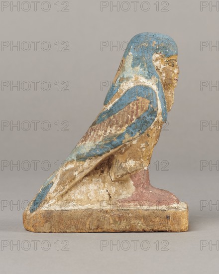 Figurine of the Soul of Ba, Ba mythology; birds, bird iconography; djed pillars iconography; wigs; Soul personification; figurines; beards facial hair; feathers. The figurine has carefully carved and painted facial details and a reproduced arrangement of feathers on the wings. On the head is a tripartite wig painted blue. On the chest below the wig is a depiction of a djed pillar. Straps supporting a false beard are painted along the cheeks. The figurine's disproportionately large feet rest on a rectangular base. 664–332 BC, Late Period, Egypt. Figurine; height 11 cm, width 9.5 cm, depth 4.5 cm., A painted wooden figure of a bird-like creature with a human face sits on a rectangular base, showing worn and chipped surfaces; the figure's head and upper body have blue paint, with areas of white and beige showing through, the face is mostly beige with traces of darker detailing, the wings display a mix of blue and reddish-brown with flaking pigments, the lower body and base show natural wo...