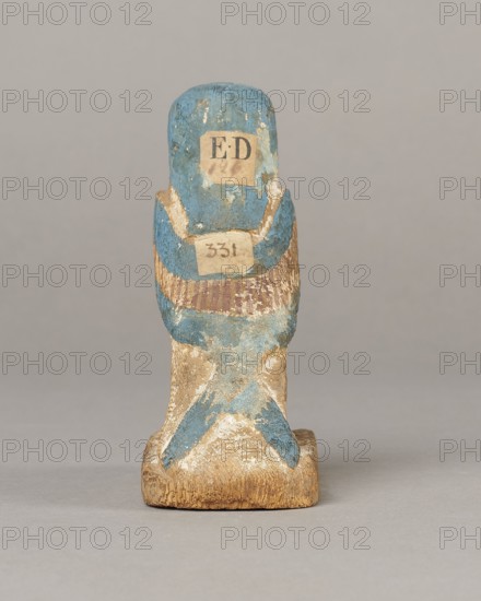 Figurine of the Soul of Ba;, Subject: Ba mythology. Description: painted and carved figurine with carefully carved and painted facial details, wings with arranged feathers, tripartite wig painted blue, depiction of a djed pillar on the chest below the wig, painted straps supporting a false beard along the cheeks, disproportionately large feet on a rectangular base. Iconography: birds, djed pillars, wigs, beards, feathers, Soul personification. Date: 664–332 BC, Late Period, Egypt. Object type: figurine. Dimensions: height 11 cm, width 9.5 cm, depth 4.5 cm., A small carved wooden figure shown from the front, with worn blue and beige paint and areas of exposed wood; the figure has a rounded top and a broader base, with traces of darker brown and off-white in places, a textured surface with chipped paint, and a neutral gray background.