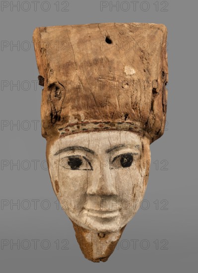 Sarcophagus mask with fragment of a high wig above the forehead, decorated on the edge of the forehead with a chain motif with details painted in black and red. The face is painted light cream; the large eyes and eyebrows are highlighted with black paint. Sarcophagus mask; masks, faces, wig iconographic; grave goods; Egyptian art; 332–30 BC, Ptolemaic Period, Egypt. Face height: 12.5 cm. Overall height 31 cm, width 17 cm, depth 9 cm., A carved wooden head with a large blocky top section showing natural wood grain and knots in brown tones, attached to a painted lower face area in pale off-white with visible cracks; the face has dark painted eyes and arched eyebrows in black, a subtle nose and mouth carved into the pale surface, and a narrow chin area where the wood is exposed in brown; a thin band of faded paint in red and green separates the wooden top from the painted face, all set against a neutral gray background.