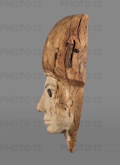 Sarcophagus mask with fragment of a high wig above the forehead, decorated on the edge of the forehead with a chain motif with details painted in black and red. The face is painted light cream; the large eyes and eyebrows are highlighted with black paint. Sarcophagus fragments; masks, faces; grave goods. Face height: 12.5 cm. Overall height 31 cm, width 17 cm, depth 9 cm. 332–30 BC; Ptolemaic Period, Egypt., A carved wooden profile of a human face shown from the side, with visible wood grain, cracks, and knots; the face area is lighter in color while the surrounding wood is a darker brown, and there are small areas of black pigment around the eye; the background is a solid gray.
