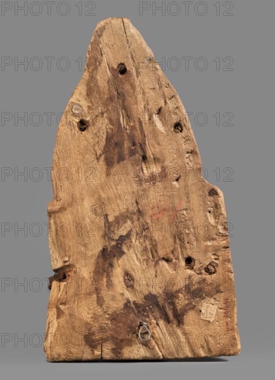 Sarcophagus mask, Sarcophagus mask with fragment of a high wig above the forehead, decorated on the edge of the forehead with a chain motif with details painted in black and red. The face is painted light cream, the large eyes and eyebrows are highlighted with black paint. Masks, faces, grave goods; wig iconographic; sarcophagus fragments. Face height: 12.5 cm. Overall height 31 cm, width 17 cm, depth 9 cm. Date: 332–30 BC, Ptolemaic Period, Egypt., A roughly triangular piece of weathered wood with an uneven, worn surface showing grain patterns, knots, and holes, resting against a flat neutral background. The wood displays a range of brown tones from light tan to darker brown stains and patches, with a few darker smudges and irregular marks across its face. There are several small circular indentations and knot holes, a small metal ring attached near the lower edge, and areas where the wood surface appears roughened or chipped. The background is a uniform gray.