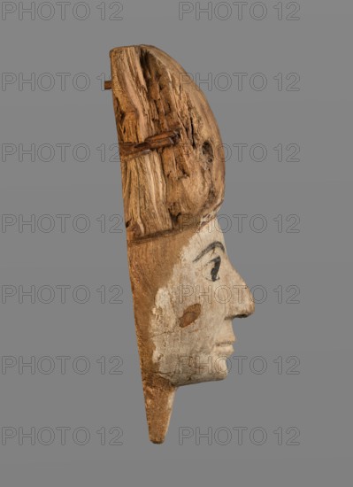 Sarcophagus mask with fragment of a high wig above the forehead, decorated on the edge of the forehead with a chain motif with details painted in black and red. The face is painted light cream; the large eyes and eyebrows are highlighted with black paint. Materials: painted wood sarcophagus mask, sarcophagus fragments. Function: funerary mask, grave goods. Style/period: Egyptian, Ptolemaic Period, 332–30 BC. Dimensions: face height 12.5 cm; overall height 31 cm, width 17 cm, depth 9 cm. Keywords: wig iconographic, masks, faces, Egyptian art., A carved wooden profile of a human face shown from the side against a plain gray background, with a weathered surface and visible wood grain and cracks; the top portion appears darker brown with rough, textured wood while the lower face area is lighter beige with remnants of pale paint and darker paint marking the eye and eyebrow; the nose and lips are rounded and the piece tapers to a pointed end below, showing a mix of brown, beige, pale off-whi...