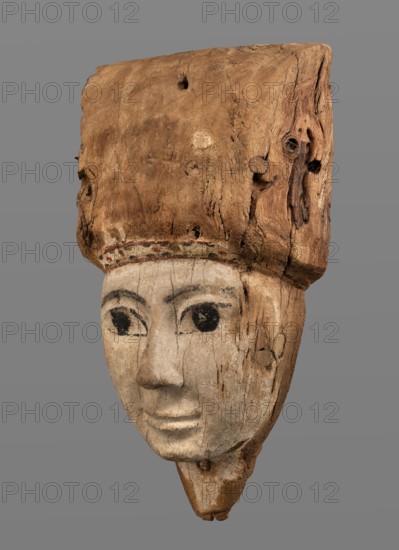 Sarcophagus mask, Sarcophagus mask with fragment of a high wig above the forehead, decorated on the edge of the forehead with a chain motif with details painted in black and red. The face is painted light cream; the large eyes and eyebrows are highlighted with black paint. Wig iconographic, sarcophagus fragments; masks, faces; grave goods. Face height: 12.5 cm. Overall height 31 cm, width 17 cm, depth 9 cm. Date: 332–30 BC, Ptolemaic Period, Egypt., A carved wooden head with a worn, light brown upper section and a pale, weathered lower face; the face shows painted dark eyes and dark eyebrow shapes, with visible cracks and grain lines across the wood; a narrow band of faint, muted color sits between the upper and lower sections; the upper wooden portion is rough, textured, and darker brown with knots and holes; the background is a uniform gray.