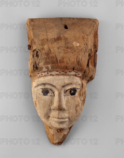 Sarcophagus mask with fragment of a high wig above the forehead, decorated on the edge of the forehead with a chain motif with details painted in black and red. The face is painted light cream; the large eyes and eyebrows are highlighted with black paint. Sarcophagus mask, wig iconographic, masks, faces; grave goods; Egyptian art; Ptolemaic Period, 332–30 BC, Egypt. Face height: 12.5 cm; overall height 31 cm, width 17 cm, depth 9 cm., A carved wooden face with a rectangular block of rough brown wood attached at the top, showing grain, knots, and a few holes; the carved face below is a lighter beige-cream color with painted black eyes and dark eyebrows, a subtle carved nose and mouth, and traces of darker coloration along the hairline where a few muted red and black spots are visible.