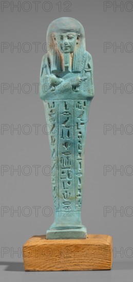 Udja-hera shabti, Udja-hera ca. 380–343 BC. Mummy-shaped funerary figurine shabti, Late Period, 30th Dynasty, Egypt. Height 15.5 cm, width 3.9 cm. Standing on a base with back supported by a pillar. Arms crossed on the chest, right folded over left; elbows and shoulders subtly marked. Holds hoes in each hand with slightly curled handles; right hand also holds a twisted rope from which a basket is slung over the left shoulder. Convex tools and an engraved basket. Wears a long three-part wig combed into narrow strands with main strands ending in borders; long beard slightly tapering and curled. Large ears; proportionate, carefully crafted facial details and a slight smile. Inscription engraved in three columns separated by lines on the front of the figurine's legs. Mentions wigs, hieroglyphs, hoes, beards, Book of the Dead, funerary figurines, grave goods., A vertical statuette in shades of turquoise and blue-green depicting a standing figure with a stylized wig and crossed arms, mounted...