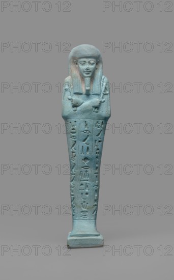 Udja-hera shabti, maker, Udja-hera ca. 380–343 BC. Mummy-shaped funerary figurine shabti, Late Period, 30th Dynasty, Egypt. Standing on a base with back supported by a pillar; height 15.5 cm, width 3.9 cm. Arms crossed on the chest right over left with subtly marked elbows and shoulders; holds hoes in each hand whose handles are slightly curled. Right hand holds a twisted rope with a basket slung over the left shoulder; convex tools and an engraved basket. Wears a long three-part wig combed into narrow strands ending in borders; long beard slightly tapering and curled; large ears; proportionate, carefully crafted facial features with a slight smile. Inscription engraved in three columns separated by lines on the front of the figurine's legs. Mentions wigs, hieroglyphs, hoes, beards, Book of the Dead, grave goods, purchase provenance., A small statuette of a standing figure with crossed arms wearing a straight wig and a narrow beard, rendered in a turquoise-blue material with subtle gre...