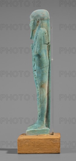 Udja-hera shabti, Udja-hera ca. 380–343 BC, wigs, hieroglyphs, hoes, beards facial hair, Book of the Dead, funerary figurine, grave goods, mummy-shaped figurine, standing on a base, back supported by a pillar. Arms crossed on the chest, right folded over left, elbows and shoulders subtly marked. Hoes in each hand; handles slightly curled. In the right hand is a twisted rope with a basket slung over the left shoulder. Convex tools, engraved basket. Long, three-part wig combed into narrow strands with main strands ending in borders. Long beard, slightly tapering and curled. Large ears. Proportionate, carefully crafted facial details; mouth set in a slight smile. Inscription engraved; arranged in three columns separated by lines on the front of the figurine's legs. 380–343 BC; Late Period, 30th Dynasty, Egypt. Shabti; height 15.5 cm, width 3.9 cm., A small turquoise-green figurine with a matte, slightly weathered surface stands upright on a rectangular wooden base; the figure is shown in ...