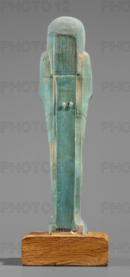 Udja-hera shabti, Udja-hera ca. 380–343 BC. Mummy-shaped funerary figurine shabti, Late Period, 30th Dynasty, Egypt. Standing on a base with back supported by a pillar; arms crossed on the chest right over left with elbows and shoulders subtly marked. Holds hoes in each hand with slightly curled handles; right hand holds a twisted rope from which a basket is slung over the left shoulder. Convex tools and engraved basket. Long three-part wig combed into narrow strands ending in borders; long slightly tapering curled beard; large ears; proportionate, carefully crafted facial details with a slight smile. Inscription engraved and arranged in three columns separated by lines on the front of the figurine's legs. Height 15.5 cm; width 3.9 cm., A weathered turquoise-green figurine stands upright on a rectangular wooden base, showing a stylized human form with a rounded head and elongated body; the surface has areas of lighter green and pale beige discoloration, small pits and abrasions, and su...