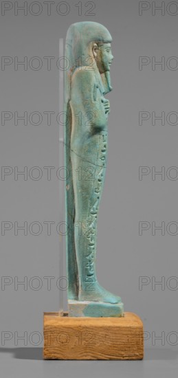 Udja-hera shabti, Udja-hera ca. 380–343 BC. Mummy-shaped funerary figurine shabti, Late Period, 30th Dynasty, Egypt. Height 15.5 cm, width 3.9 cm. Standing on a base with back supported by a pillar; arms crossed on the chest right over left with elbows and shoulders subtly marked. Holds hoes in each hand with slightly curled handles; right hand holds a twisted rope from which a basket is slung over the left shoulder. Convex tools and an engraved basket depicted. Wears a long three-part wig combed into narrow strands ending in borders; long slightly tapering, curled beard; large ears; proportionate, carefully crafted facial features and a slight smile. Inscription engraved in three columns separated by lines on the front of the legs. Materials/attributes: wigs, hieroglyphs, hoes, beards; function as grave goods linked to the Book of the Dead. Provenance: purchase., A small standing figure sculpture shown in profile on a wooden base, the figure is a greenish-blue with weathered lighter a...