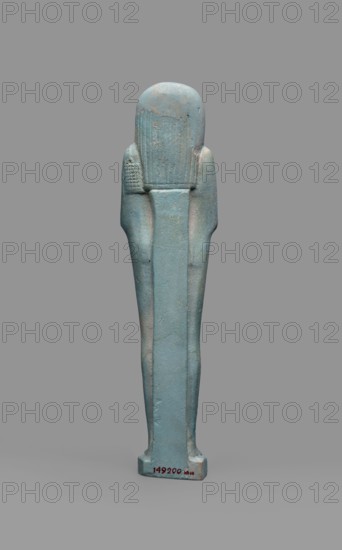 Udja-hera shabti, Udja-hera ca. 380–343 BC. Mummy-shaped funerary figurine shabti, Late Period, 30th Dynasty, Egypt. Height 15.5 cm, width 3.9 cm. Standing on a base with back supported by a pillar. Arms crossed on the chest, right folded over left; elbows and shoulders subtly marked. Holds hoes in each hand with slightly curled handles; right hand also holds a twisted rope from which a basket is slung over the left shoulder. Convex tools and an engraved basket. Long three-part wig combed into narrow strands ending in borders; long slightly tapering curled beard; large ears; proportionate, carefully crafted facial details with a slight smile. Inscription engraved in three columns separated by lines on the front of the legs. Mentions wigs, hieroglyphs, hoes, beards, Book of the Dead, funerary figurines, grave goods., A tall, vertical statuette with a smooth, elongated form in shades of blue and turquoise, showing areas of pale greenish-blue and subtle beige wear; the figure has a rounde...
