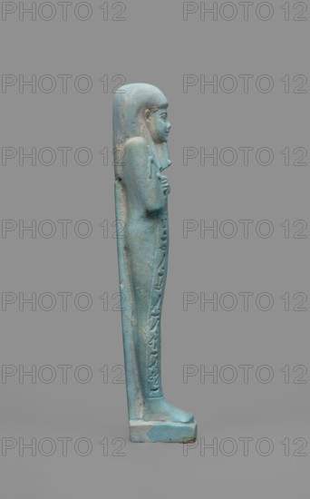 Udja-hera shabti, Udja-hera ca. 380–343 BC. Mummy-shaped funerary figurine shabti, Late Period, 30th Dynasty, Egypt. Height 15.5 cm; width 3.9 cm. Standing on a base with back supported by a pillar. Arms crossed on the chest right folded over left, elbows and shoulders subtly marked. Holds hoes in each hand; handles slightly curled. Right hand holds a twisted rope with a basket slung over the left shoulder. Convex tools and an engraved basket. Long three-part wig combed into narrow strands with bordered main strands. Long beard, slightly tapering and curled. Large ears; proportionate, carefully crafted facial details and a slight smile. Inscription engraved in three columns separated by lines on the front of the figurine's legs. Mentions of wigs, hieroglyphs, hoes, beards facial hair, Book of the Dead, funerary figurines, grave goods., A vertical turquoise-blue statuette of a standing human figure shown in profile against a solid gray background, with long hair or a headdress that exte...