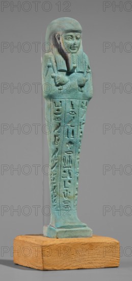 Udja-hera shabti, Udja-hera ca. 380–343 BC. Mummy-shaped funerary figurine shabti, Late Period, 30th Dynasty, Egypt. Height 15.5 cm; width 3.9 cm. Standing on a base with back supported by a pillar. Arms crossed on the chest right folded over left, elbows and shoulders subtly marked. Holds hoes in each hand with slightly curled handles; right hand also holds a twisted rope from which a basket is slung over the left shoulder. Convex tools and engraved basket. Wears a long three-part wig combed into narrow strands with main strands ending in borders; long tapering, curled beard; large ears; proportionate, carefully crafted facial details with a slight smile. Inscription engraved in three columns separated by lines on the front of the figurine's legs. Mentions of wigs, hieroglyphs, hoes, beards, Book of the Dead, grave goods, and funerary function., A small carved figurine stands on a rectangular wooden base; the figurine is turquoise and bluish-green with areas of darker green and slight...