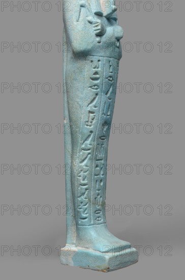 Udja-hera shabti, Udja-hera ca. 380–343 BC. Mummy-shaped funerary figurine shabti, Late Period, 30th Dynasty, Egypt. Height 15.5 cm; width 3.9 cm. Standing on a base, back supported by a pillar. Arms crossed on the chest, right folded over left; elbows and shoulders subtly modeled. Holds hoes in each hand with slightly curled handles. Right hand holds a twisted rope from which a basket is slung over the left shoulder; convex tools and engraved basket. Long three-part wig combed into narrow strands with main strands ending in borders. Long, slightly tapering curled beard; large ears; proportionate, carefully crafted facial details with a slight smile. Inscription engraved in three columns separated by lines on the front of the figurine's legs. Mentions wigs, hieroglyphs, hoes, beards, Book of the Dead, funerary figurines, grave goods., A turquoise-blue elongated figurine fragment showing the lower half of a standing human form with a few carved vertical grooves and a column of raised ca...