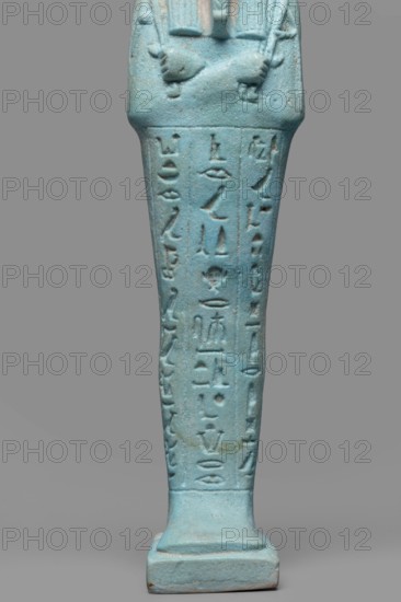 Udja-hera shabti, Udja-hera ca. 380–343 BC. Late Period, 30th Dynasty, Egypt. Mummy-shaped funerary figurine shabti, standing on a base with back supported by a pillar; height 15.5 cm, width 3.9 cm. Arms crossed on the chest, right over left, elbows and shoulders subtly marked. Holds hoes in each hand with slightly curled handles; right hand holds a twisted rope with a basket slung over the left shoulder. Convex tools and an engraved basket. Wears a long three-part wig combed into narrow strands ending in borders, and a long beard tapering and curled. Large ears; proportionate, carefully crafted facial details with a slight smile. Inscription engraved in three columns separated by lines on the front of the figurine's legs. Provenance: grave goods; related to Book of the Dead motifs., A close-up of a vertical turquoise-blue statuette fragment showing the lower torso, crossed arms, and elongated legs on a rectangular base against a gray background; the surface has incised vertical groove...