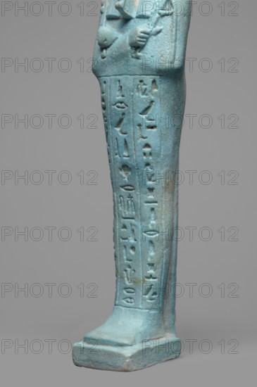 Udja-hera shabti, Udja-hera ca. 380–343 BC. Mummy-shaped funerary figurine shabti, Late Period, 30th Dynasty, Egypt. Height 15.5 cm, width 3.9 cm. Standing on a base with back supported by a pillar. Arms crossed on the chest, right folded over left; elbows and shoulders subtly marked. Holds hoes in each hand with slightly curled handles; twisted rope in right hand suspending a basket over the left shoulder. Convex tools and an engraved basket depicted. Wearing a long three-part wig combed into narrow strands with bordered ends; long slightly tapering curled beard; large ears; proportionate facial details with a slight smile. Inscription engraved in three columns separated by lines on the front of the legs. Associated motifs/attributes: wigs, hieroglyphs, hoes, beards, Book of the Dead; function as grave goods/funerary figurine. Purchase, A close-up of the lower portion of a standing figurine rendered in a turquoise-blue material, showing a pair of crossed arms at the top of the frame a...