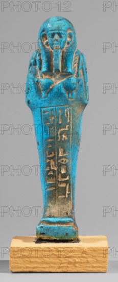 Padiamonnesut's shabti, Padiamonnesut ca. 664–525 BC, Tadiasetmen ca. 664–525 BC. Mummy-shaped figurine, standing on a base, supported at the back on a pillar. Hands crossed on the chest, the right folded over the left. In the left hand a pickaxe, in the right a hoe, and a twisted string with a basket slung over the left shoulder. Smooth three-part wig, large ears, slightly closed eyes, mouth in a slight smile, long straight beard. Engraved inscription in two columns in a frame on the front of the figurine's legs. 664–525 BC; Late Period, 26th Dynasty, Egypt. Shabti; height 11.2 cm, width 3.2 cm., A small blue-green figurine shaped like a standing mummiform figure is shown on a rectangular yellow-beige base; the figure has a rounded head with a stylized wig or headdress, a visible face with eye and mouth contours, crossed arms over the chest, and a long tapered body with shallow vertical recessed lines and some incised vertical markings that are visually distinct from the surrounding s...