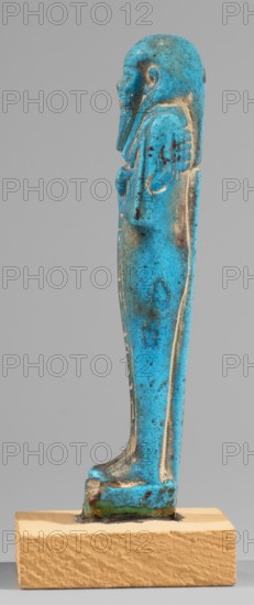 Padiamonnesut's shabti, Padiamonnesut ca. 664–525 BC, Tadiasetmen ca. 664–525 BC. Mummy-shaped figurine shabti, Late Period, 26th Dynasty, Egypt; height 11.2 cm, width 3.2 cm. Standing on a base, supported at the back by a pillar. Hands crossed on the chest, right folded over left; left hand holds a pickaxe, right a hoe, with a twisted string carrying a basket over the left shoulder. Smooth three-part wig, large ears, slightly closed eyes, slight smile, long straight beard. Engraved inscription in two columns in a frame on the front of the figurine's legs., A small upright figurine rendered in bright turquoise blue with areas of darker blue and small patches of brownish discoloration, standing on a rectangular beige base; the figure has a rounded head, a carved facial profile and a long beard, with arms folded or held close to the body and a long robe that tapers down to flat feet, and the surface shows slight pitting and worn edges where an underlying lighter or off-white material is ...