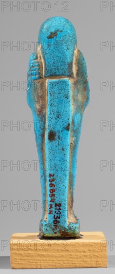 Padiamonnesut's shabti, Padiamonnesut ca. 664–525 BC, Tadiasetmen ca. 664–525 BC. Mummy-shaped figurine, standing on a base, supported at the back by a pillar. Hands crossed on the chest, right folded over left; left hand holds a pickaxe, right a hoe, and a twisted string with a basket slung over the left shoulder. Smooth three-part wig, large ears, slightly closed eyes, mouth in a slight smile, long straight beard. Engraved inscription in two columns in a frame on the front of the figurine's legs. 664–525 BC; Late Period, 26th Dynasty, Egypt. Shabti. Height 11.2 cm; width 3.2 cm., A small figurine shaped like a standing human form with arms at its sides, shown from the back, with a smooth rounded head and a long narrow body tapering toward the base, mounted on a rectangular block. The figurine is predominantly bright turquoise blue with areas of darker blue speckling and scattered brownish spots and patches, and has worn pale beige or tan exposed areas along raised edges and around th...