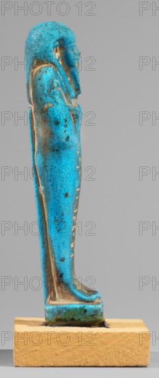Padiamonnesut's shabti. Mummy-shaped figurine, standing on a base, supported at the back on a pillar. Hands crossed on the chest, right folded over left. Holds a pickaxe in the left hand and a hoe in the right; a twisted string with a basket is slung over the left shoulder. Wears a smooth three-part wig; large ears; eyes slightly closed; mouth in a slight smile; long straight beard. Engraved inscription in two columns in a frame on the front of the legs. 664–525 BC; Late Period, 26th Dynasty, Egypt. Shabti; height 11.2 cm, width 3.2 cm., A slender vertical figurine in bright turquoise blue with mottled darker blue and brown spots stands on a rectangular beige base, the figure showing a human profile with a headdress and a long wrapped garment, traces of a lighter worn color along some edges, subtle shadows and highlights on the curved surfaces, and a plain pale gray background.
