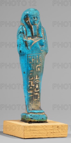 Padiamonnesut's shabti. Mummy-shaped figurine, standing on a base, supported at the back on a pillar. Hands crossed on the chest, right folded over left. Left hand holds a pickaxe, right a hoe; a twisted string with a basket slung over the left shoulder. Smooth three-part wig, large ears, slightly closed eyes, mouth in a slight smile, long straight beard. Engraved inscription in two columns framed on the front of the figurine's legs. 664–525 BC; Late Period, 26th Dynasty, Egypt. Shabti; height 11.2 cm, width 3.2 cm., A small statue of a standing human figure with folded arms, shown against a plain gray background, with a few visible areas of surface wear and pitting; the figure has a stylized headdress and a calm facial expression. The statue is predominantly bright turquoise blue with patches of darker blue and areas where a sandy beige or tan color shows through from worn or exposed material. The base supporting the statue is a rough-textured rectangular block in a warm yellow-beige ...
