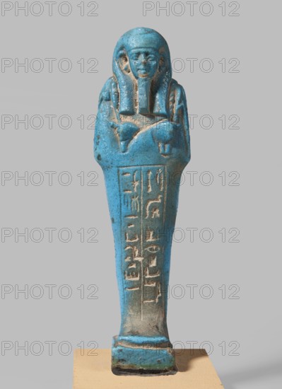 Padiamonnesut's shabti; Padiamonnesut ca. 664–525 BC, Tadiasetmen ca. 664–525 BC. Mummy-shaped figurine shabti, Late Period, 26th Dynasty, Egypt, 664–525 BC. Standing on a base, supported at the back by a pillar. Hands crossed on the chest, right folded over left; left hand holds a pickaxe, right a hoe, and a twisted string with a basket slung over the left shoulder. Smooth three-part wig, large ears, slightly closed eyes, slight smile, long straight beard. Engraved inscription in two columns within a frame on the front of the legs. Height 11.2 cm; width 3.2 cm., A blue-green statuette of a standing figure with a rounded head and a headdress, arms crossed over the chest, and a long tapering body mounted on a small rectangular base; the surface shows patches of worn beige and darker brown where the blue-green has eroded, and the background is a flat light gray.