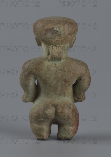 Amulet - Ptah-dwarf Pataikos, Ptah-dwarf mythology. Figurine of a naked dwarf man with a large head with a wide forehead, short arms with clenched fists pressed to the hips, and short legs broken off at the knees. Careful modelling. 7th–4th century BC; Late Period, Egypt. Figurine, amulet. Height 6.5 cm; width 3.7 cm; depth 2.6 cm., A small sculpted figure shown from the back standing on a flat surface against a plain gray background; the figure has a rounded head, short neck, broad shoulders, bent arms resting at the sides, a defined back and buttocks, and short legs with a gap between them. The surface of the figure shows a weathered, mottled patina with shades of green, brown, and hints of blue, and a small area with a faded red tint. The texture appears rough and aged with areas of darker and lighter discoloration.
