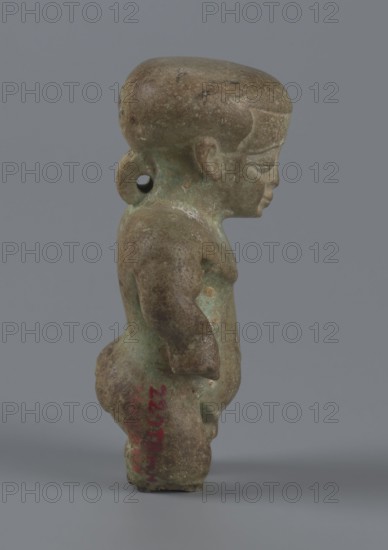 Amulet — Ptah-dwarf Pataikos, Figurine of a naked dwarf man with a large head with a wide forehead, short arms with clenched fists pressed to the hips, and short legs broken off at the knees. Carefully modelled. Ptah-dwarf mythology. 7th–4th century BC, Late Period, Egypt. Figurine/amulet. Height 6.5 cm; width 3.7 cm; depth 2.6 cm., A small figurine viewed in profile with a rounded head and a pronounced forehead, showing a facial profile with a closed mouth and a softly defined nose; the body is compact with rounded limbs and a slightly protruding belly, and there is a loop-like projection at the back of the head with a hole through it. The surface shows a mottled patina in shades of green and brown with areas of darker brown and some lighter greenish spots, and a cool gray background behind the figure.