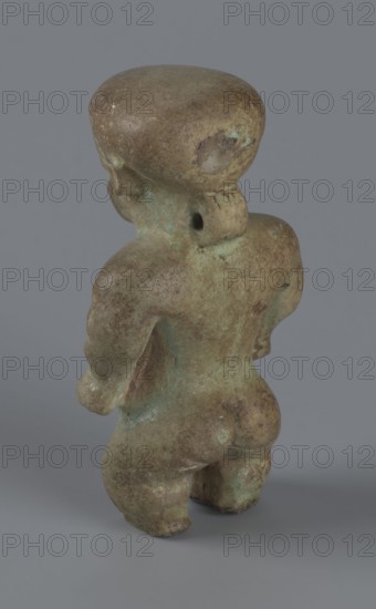 Amulet - Ptah-dwarf Pataikos, Ptah-dwarf mythology. Figurine of a naked dwarf man with a large head with a wide forehead, short arms with clenched fists pressed to the hips, and short legs broken off at the knees. Careful modelling. 7th–4th century BC; Late Period, Egypt. Figurine, amulet. Height 6.5 cm, width 3.7 cm, depth 2.6 cm., A small, weathered figurine shown from the back standing on a flat surface, with a rounded head, short neck, broad shoulders, and a rounded torso and legs; the surface shows an uneven, mottled texture with patches of greenish patina and areas of brown and tan discoloration, and a few darker spots; the background is a smooth, neutral gray.