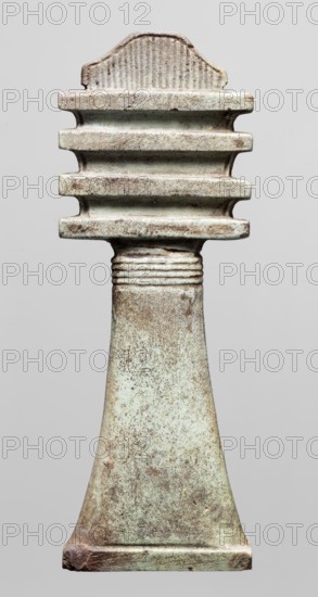 Amulet - djed pillar. Amulet in the shape of the djed hieroglyph, provided with a pillar at the back. At the top a trapezoidal projection decorated with vertical striations, below four crossbars. The top of the shaft provided with horizontal incisions. Protective amulet used in grave equipment. Late Period, Egypt, 664–332 BC. Amulet; height 9 cm, width 3.3 cm., An upright carved object with a tapered rectangular shaft and a stepped top, showing a textured surface with a patina of pale green and brown; the top has a series of horizontal ridges and a rounded crest with vertical striations, the neck area displays several narrow encircling grooves, and the base flares slightly with worn edges and mottled spotting in shades of green, brown, and beige.
