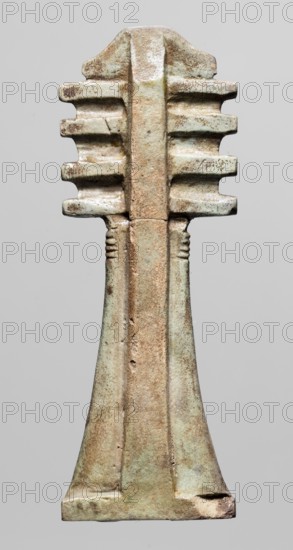 Amulet — djed pillar. Amulet in the shape of the djed hieroglyph with a pillar at the back; at the top a trapezoidal projection decorated with vertical striations and below four crossbars; the top of the shaft has horizontal incisions. Material: amulet; period: Late Period, Egypt, 664–332 BC. Dimensions: height 9 cm, width 3.3 cm., A vertical carved object with a symmetrical form, showing a textured surface in shades of pale green, light brown, beige, and hints of darker brown; the top has a rounded central ridge with tiered horizontal protrusions on both sides that create a stacked, ribbed appearance; the middle narrows into a slender, slightly raised central column with subtle linear grooves near its upper portion; the lower section flares outward into a trapezoidal base with worn and rough edges, exhibiting variations in patina and small surface pits and scratches.