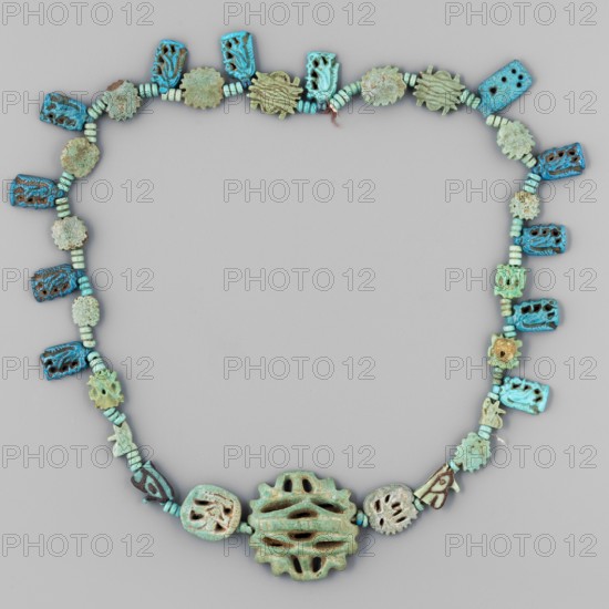 Necklace of 33 amulets and plaques, separated by tiny spherical beads several, usually 3 in a row, including green and blue faience. 12 openwork plaques with Toeris 1.2 x 0.7 cm; 1 openwork quadruple udjat eye 2.5 x 2.5 cm; 2 openwork udjat eyes 1 x 1 cm; 2 quadruple udjat eyes 1 x 1 cm; several single eyes and plates. Date: 664–332 BC Late Period, Egypt. Jewelry amulet, height 38 cm., A necklace made of carved beads arranged in a loop on a neutral gray background, featuring a range of turquoise and green hues with areas of brownish patina and pale off-white weathering; the beads include several rectangular carved pieces with openwork patterns, a few small cylindrical spacer beads, and a variety of irregularly shaped floral- or gear-like beads, with a larger central carved pendant that is rounded with slotted openings and a textured surface showing turquoise, green, and brown tones.