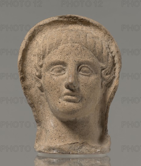 Votive head, Head of a young man covered with a shawl, from under which hair emerges in the form of a fringe and falls in curls below the temples, covering the ears. Large, wide-open, convex eyes; large, straight nose; narrow mouth with wide, slightly curled, pouting lips. Face with a strong chin alluding to classical traditions. 4th century BC, Central Italy; votive offering. Height 27.3 cm, width 20.5 cm, depth 18.5 cm, weight 3.5 kg., A weathered stone head sculpture shown against a plain gray background, featuring a rounded halo-like rim around the top and sides of the head, stylized short hair carved above the forehead, almond-shaped eyes without detailed pupils, a defined nose and closed lips, and carved ears partly visible beneath the hair; the surface shows a grainy texture with small dark speckles and areas of minor chipping and erosion. The visible colors are tan, beige, light brown, dark gray speckles, and a neutral gray background.