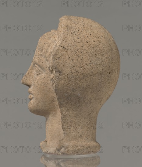 Votive head, Head of a young man covered with a shawl, from under which hair emerges in the form of a fringe and falls in curls below the temples, covering the ears. Large, wide-open, and convex eyes, large, straight nose, narrow mouth with wide, slightly curled, as if pouting lips. The face with a strong chin alludes to classical traditions. 4th century BC, Central Italy, votive offering; height 27.3 cm, width 20.5 cm, depth 18.5 cm, weight 3.5 kg., A sculpted stone head in profile facing left, showing a smooth rounded skull and defined facial features including a straight nose, closed lips, and a subtle chin, with a visible ear and traces of carved hair near the temple; the surface is textured and pitted with a grainy appearance, exhibiting a warm tan color with darker gray-brown speckles and small areas of lighter beige, set against a neutral gray background and resting on a clear circular base that reflects slightly.