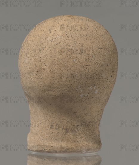 Votive head, unknown creator, Head of a young man covered with a shawl, from under which hair emerges in the form of a fringe and falls in curls below the temples, covering the ears. Large, wide-open, and convex eyes, large, straight nose, narrow mouth with wide, slightly curled, as if pouting lips. The face with a strong chin alludes to classical traditions. 4th century BC, Central Italy, votive offering. Height 27.3 cm, width 20.5 cm, depth 18.5 cm, weight 3.5 kg., A rounded, head-shaped object with a textured, porous surface in sandy beige and light brown tones sits against a solid gray background, showing small darker speckles and a few pale whitish spots on its surface; the base widens slightly and rests on a reflective surface that mirrors the bottom edge, and subtle shadows and variations in beige and brown indicate uneven texture across the form.