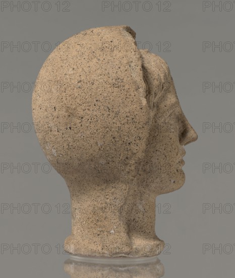 Votive head, Head of a young man covered with a shawl, from under which hair emerges in the form of a fringe and falls in curls below the temples, covering the ears. Large, wide-open, and convex eyes, large, straight nose, narrow mouth with wide, slightly curled, as if pouting lips. The face with a strong chin alludes to classical traditions. 4th century BC, Central Italy, votive offering. Height 27.3 cm, width 20.5 cm, depth 18.5 cm, weight 3.5 kg., A side view of a sculpture of a head made of a coarse beige stone with darker speckles and small lighter flecks, showing a smooth rounded back, a defined nose and lips, a subtle chin, and a rounded neck that rests on a short pedestal; the background is a flat gray and the base shows a reflection on a glossy surface.