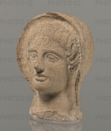 Votive head, Head of a young man covered with a shawl, from under which hair emerges in the form of a fringe and falls in curls below the temples, covering the ears. Large, wide-open, convex eyes; large, straight nose; narrow mouth with wide, slightly curled, as if pouting lips. The face with a strong chin alludes to classical traditions. 4th century BC, Central Italy, votive offering. Height 27.3 cm, width 20.5 cm, depth 18.5 cm, weight 3.5 kg., A carved stone head with a rounded top and a broken edge on one side sits against a plain gray background; the surface is sandy tan with darker speckling and small lighter flecks, showing carved facial features including shallow eye sockets, a defined nose with some surface damage at the tip, softly contoured lips, and a smooth neck that rests on a flat base; hair is indicated by shallow grooves and a few textured curls at the temple, and the stone shows areas of roughness and small pits throughout, with the whole object reflected faintly on t...