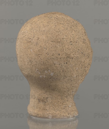 Votive head, Head of a young man covered with a shawl, from under which hair emerges in the form of a fringe and falls in curls below the temples, covering the ears. Large, wide-open, convex eyes; large, straight nose; narrow mouth with wide, slightly curled, as if pouting lips. The face with a strong chin alludes to classical traditions. 4th century BC, Central Italy, votive offering. Height 27.3 cm, width 20.5 cm, depth 18.5 cm, weight 3.5 kg., The image shows the back of a rounded, textured object resembling a head or bulbous form, with a rough, porous surface in tan and beige tones with small darker speckles and a few lighter flecks; the base narrows into a neck-like section that sits on a translucent, slightly reflective support, and the background is a uniform gray.